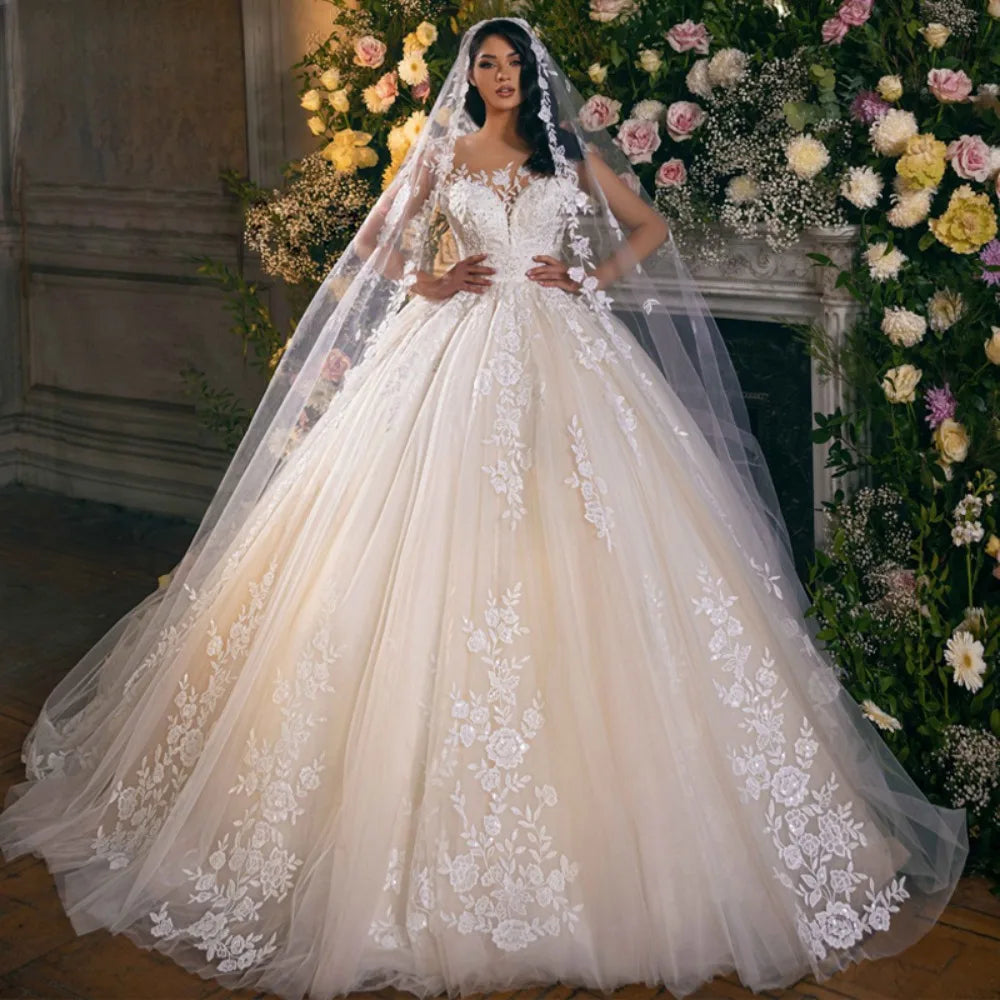 Bride in a voluminous ivory wedding gown adorned with lace floral patterns. She stands in front of a floral backdrop, wearing a long veil and posing elegantly.