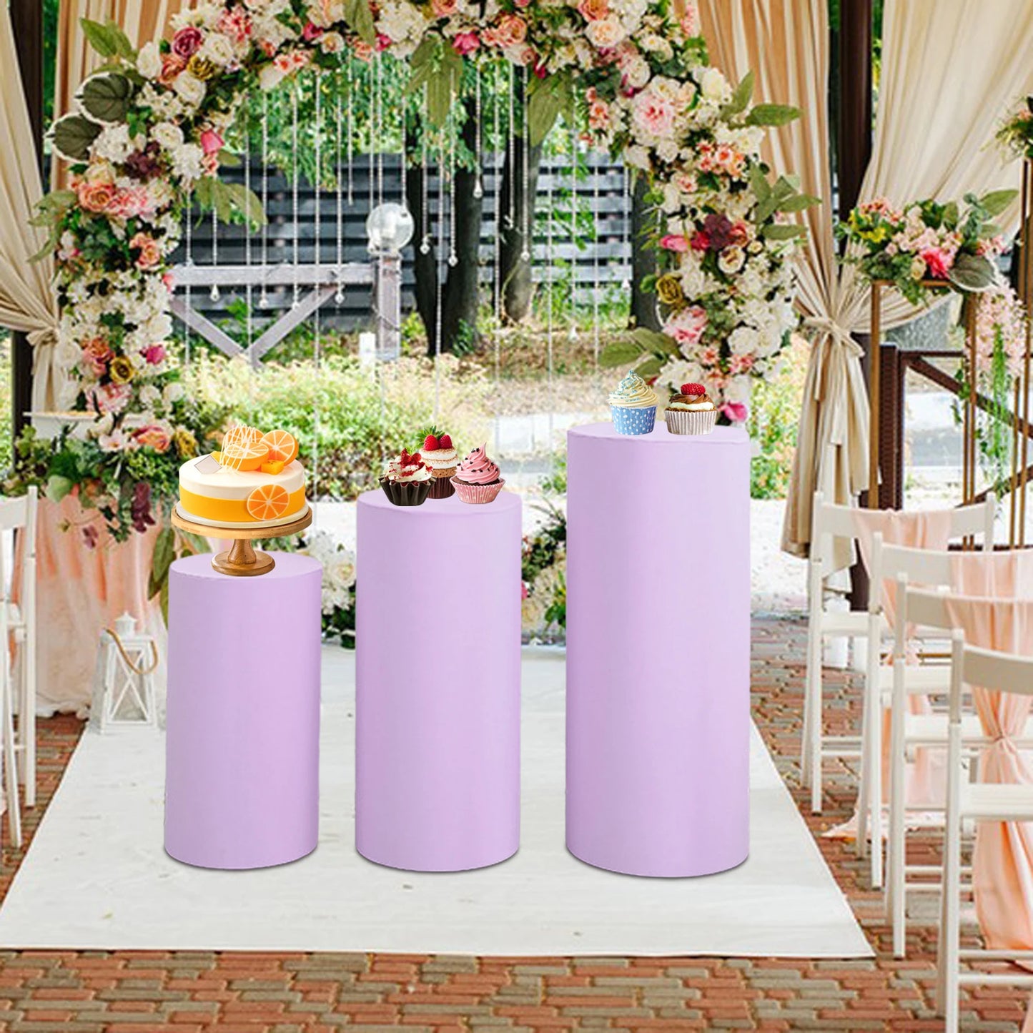 Lavender pedestals display a cake and cupcakes under a floral arch at an outdoor wedding venue. White chairs and draped fabric enhance the elegant setting.