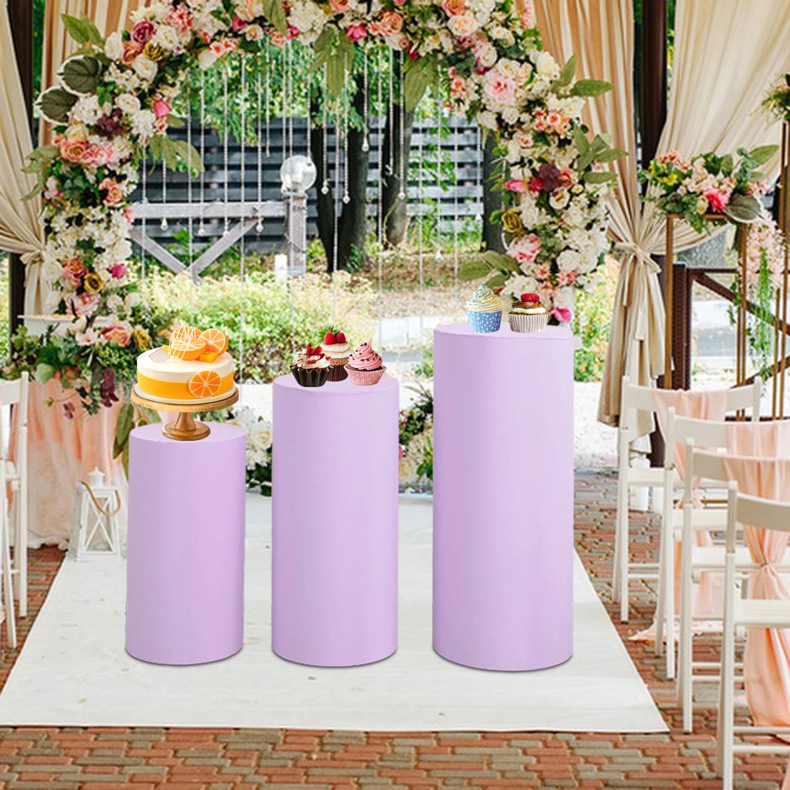 Lavender pedestals display a cake and cupcakes under a floral arch at an outdoor wedding venue. White chairs and draped fabric enhance the elegant setting.