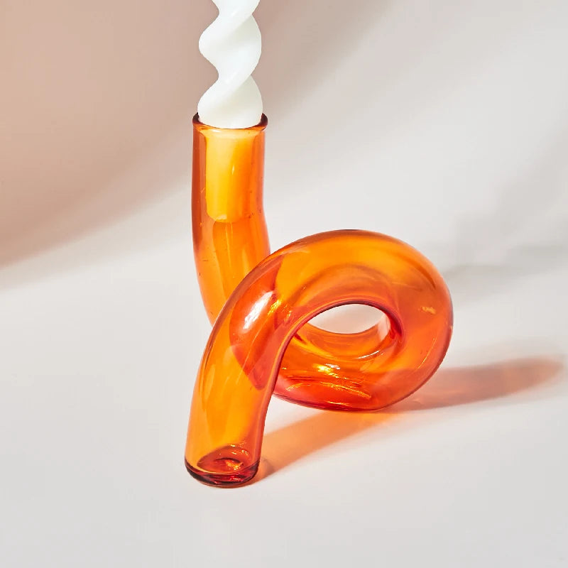 Curved orange glass candle holder with a modern design holds a white spiral candle. The holder's glossy surface casts soft shadows on the light background.