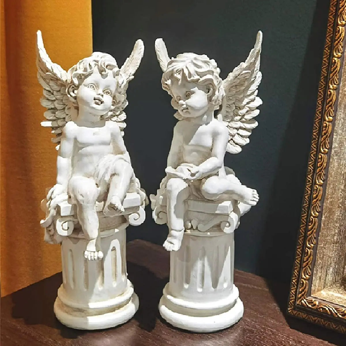 Two white angel sculptures with detailed wings are perched on ornate columns. They sit on a wooden surface against a dark green background, framed by a decorative picture frame.