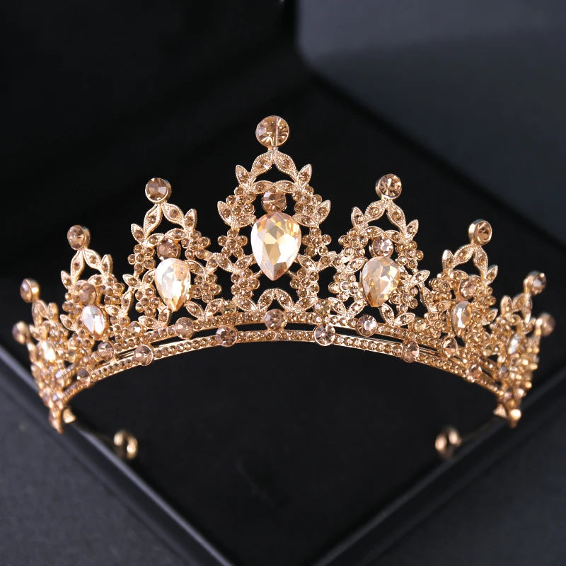 Gold tiara adorned with intricate floral patterns, sparkling gemstones, and pear-shaped crystals. The tiara is elegantly set against a dark background.