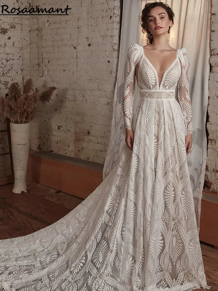 Woman in a detailed, lace wedding dress with long sleeves stands in a rustic room with a brick wall and pampas grass in a vase. The gown features a plunging neckline and a flowing train.