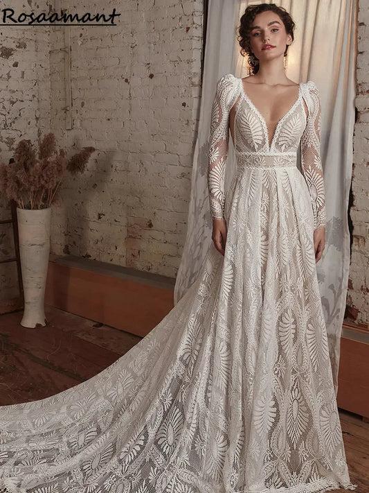 Woman in a detailed, lace wedding dress with long sleeves stands in a rustic room with a brick wall and pampas grass in a vase. The gown features a plunging neckline and a flowing train.