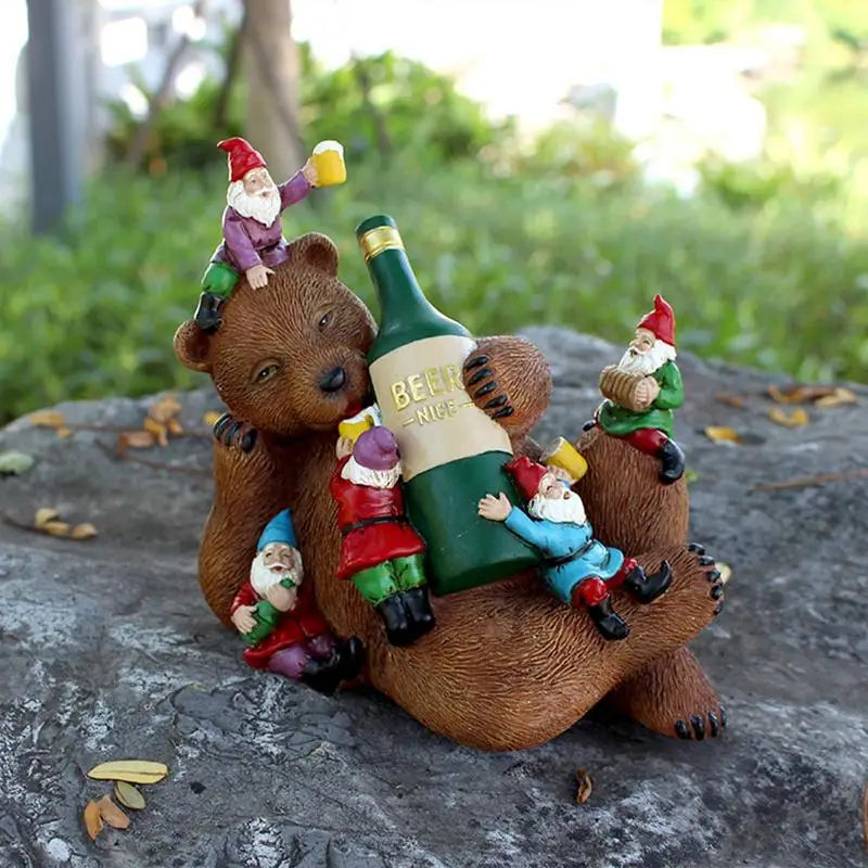 A bear figurine holds a large beer bottle while five colorful gnome figurines climb around it, enjoying a playful scene on a rock outdoors with greenery in the background.