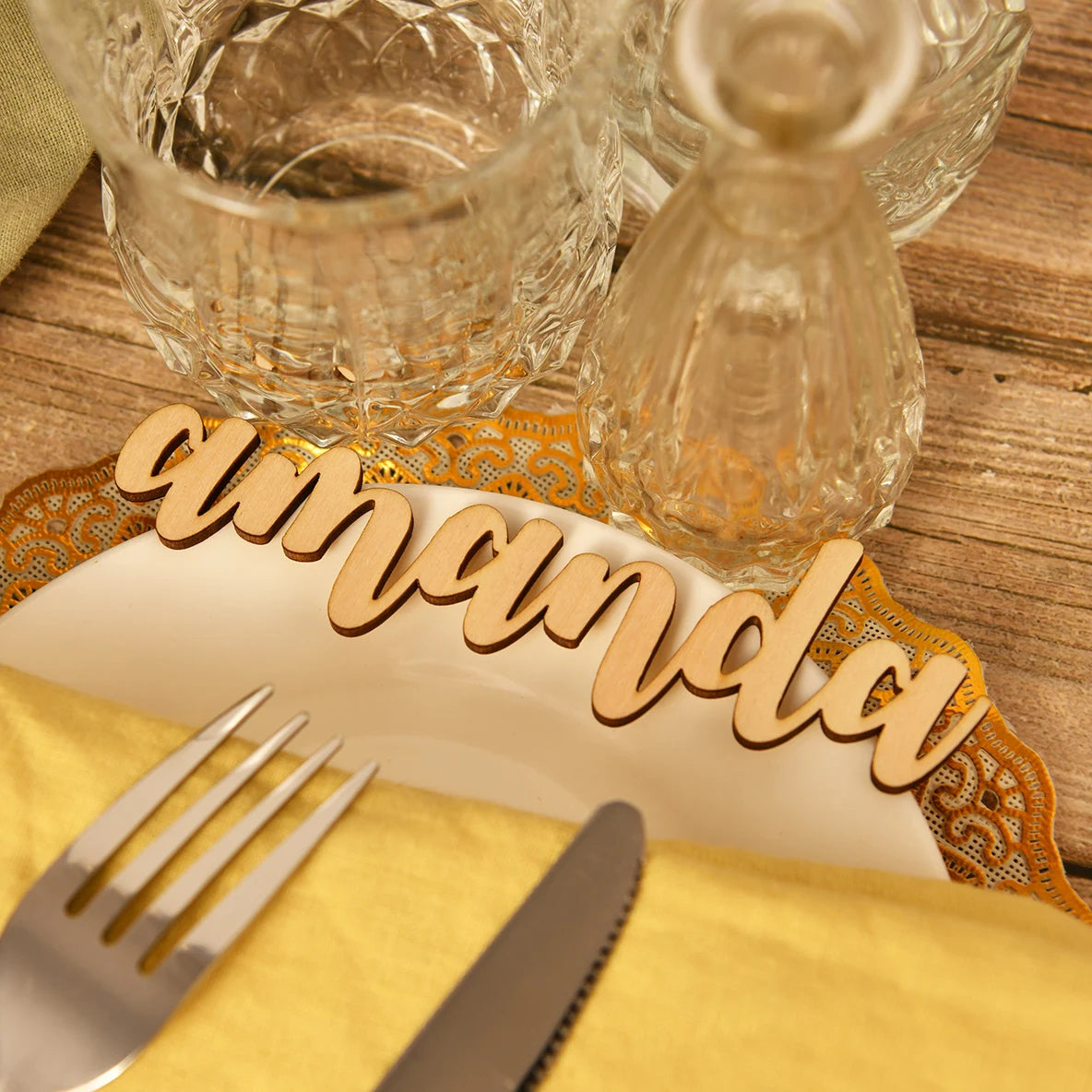 Elegant table setting with a wooden name place card on a white plate, ornate gold details, crystal glassware, and silver cutlery on a yellow napkin.