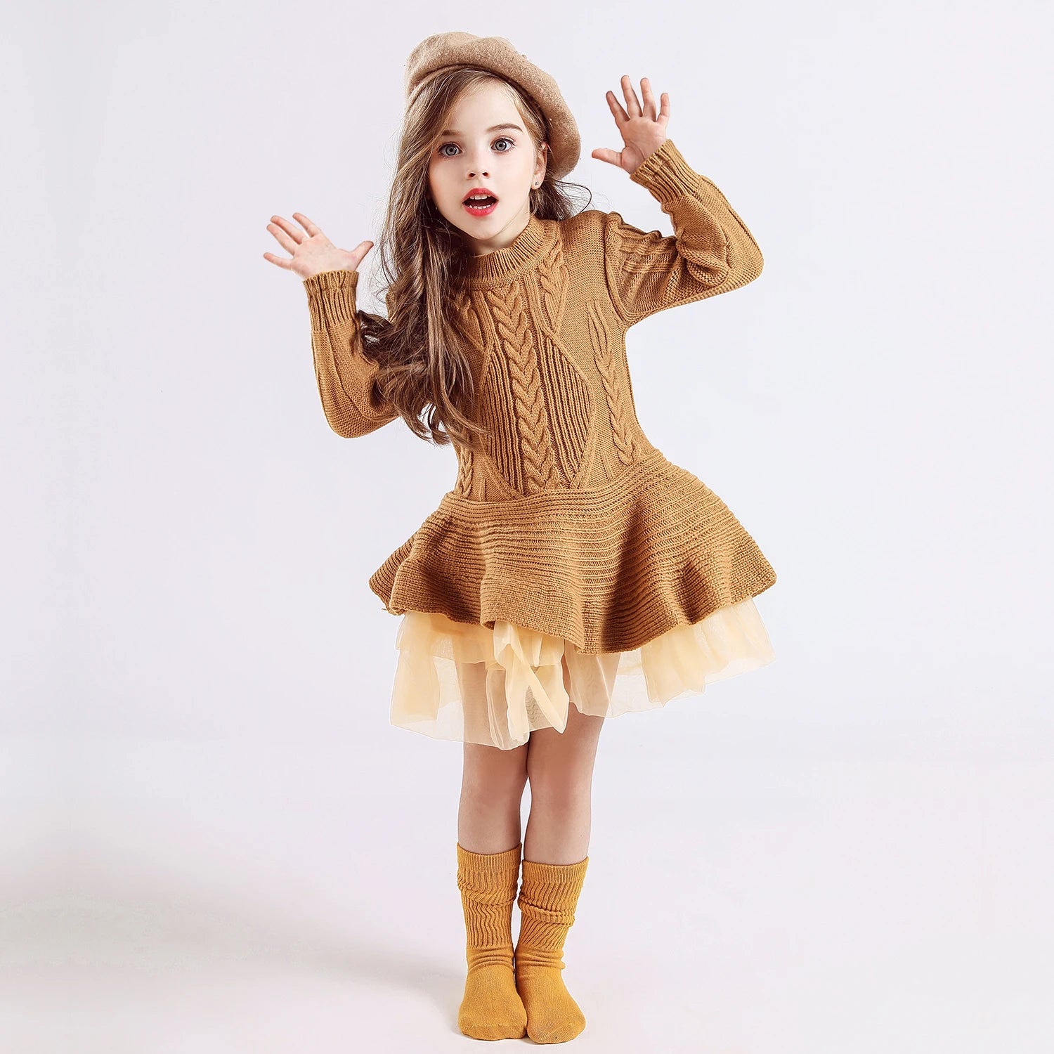 A young girl wearing a brown knit dress with a flared skirt and a beige beret poses with surprised expression, hands up. She has matching brown socks and loose wavy hair.