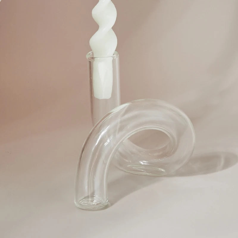 A twisted white candle in a clear, straight holder is beside a curved, transparent glass tube on a soft beige background.
