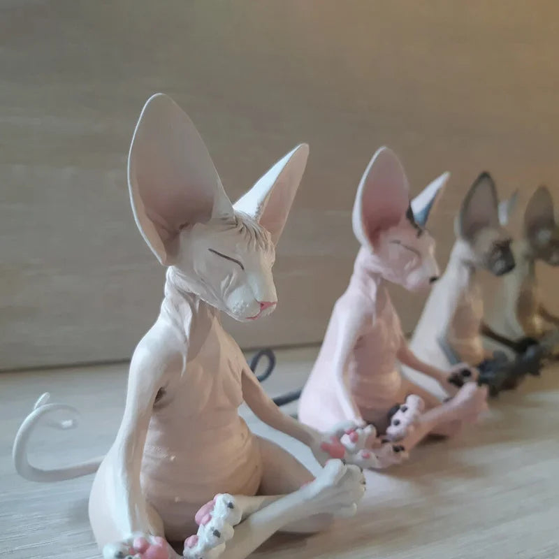 Clay sculptures of anthropomorphic hairless cats in a meditative pose sit in a row on a wooden surface, each with large ears, closed eyes, and paws resting on their knees.