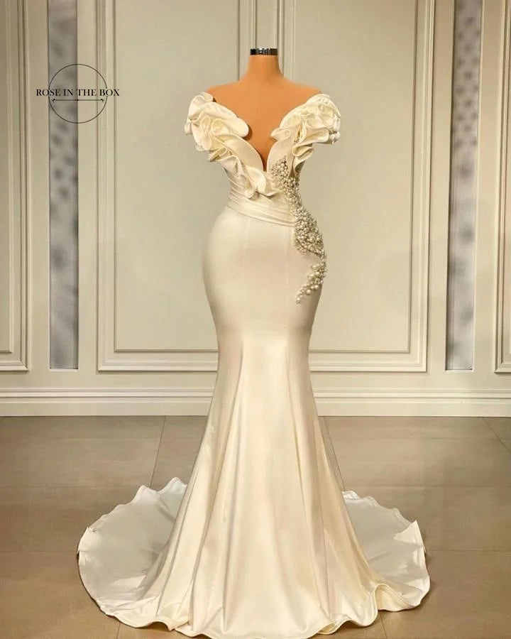 Elegant mermaid-style ivory gown on mannequin with ruffled off-shoulder sleeves and pearl detailing on the bodice, set against a white paneled wall.