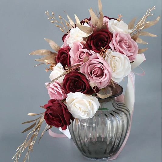 A bouquet of maroon, pink, and white roses with gold leaves arranged in a translucent, ribbed vase against a gray background.
