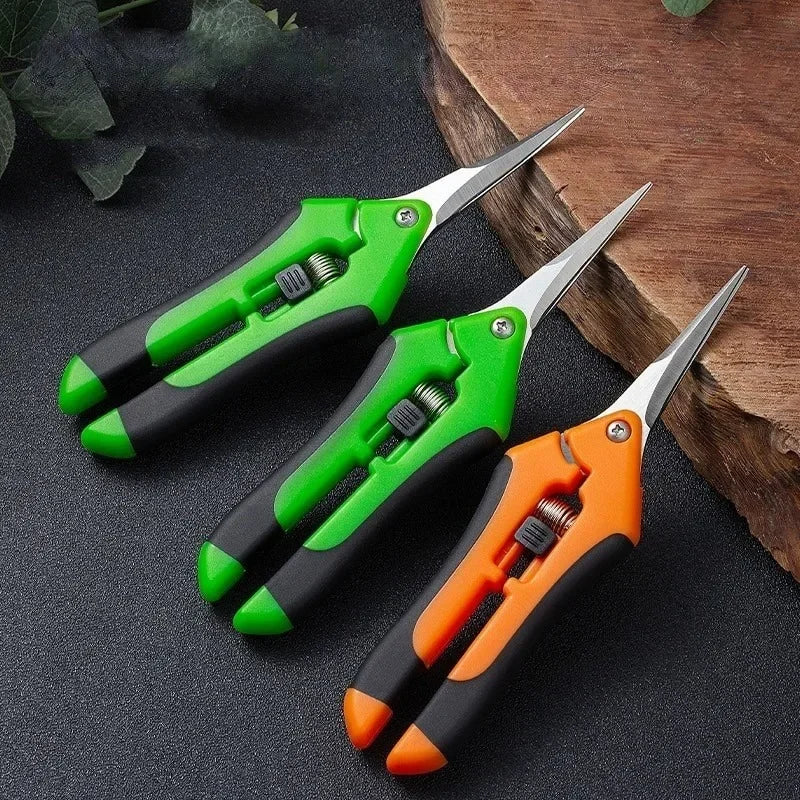 Three pairs of pruning shears with green and orange handles are arranged neatly on a dark textured surface next to a wooden slab.