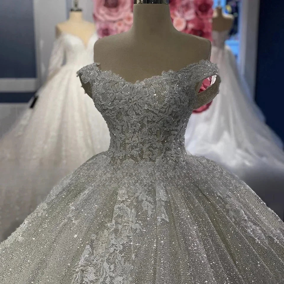 Close-up of a sparkling, off-shoulder wedding gown with intricate lace bodice on a mannequin. A floral backdrop and another elegant dress are visible in the background.