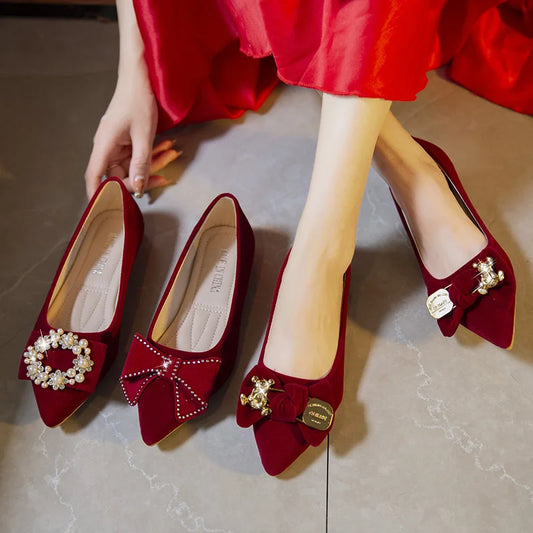 A person wearing a red dress is trying on red velvet flats with decorative bows and embellishments on a gray tile floor, showing three different shoe designs.