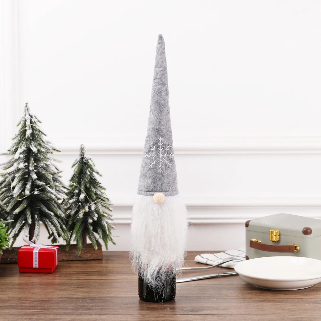 Gnome wine bottle cover with a tall gray hat, white beard, and snowflake design, set on a wooden table with small snowy trees, a gift box, and dishware in the background.