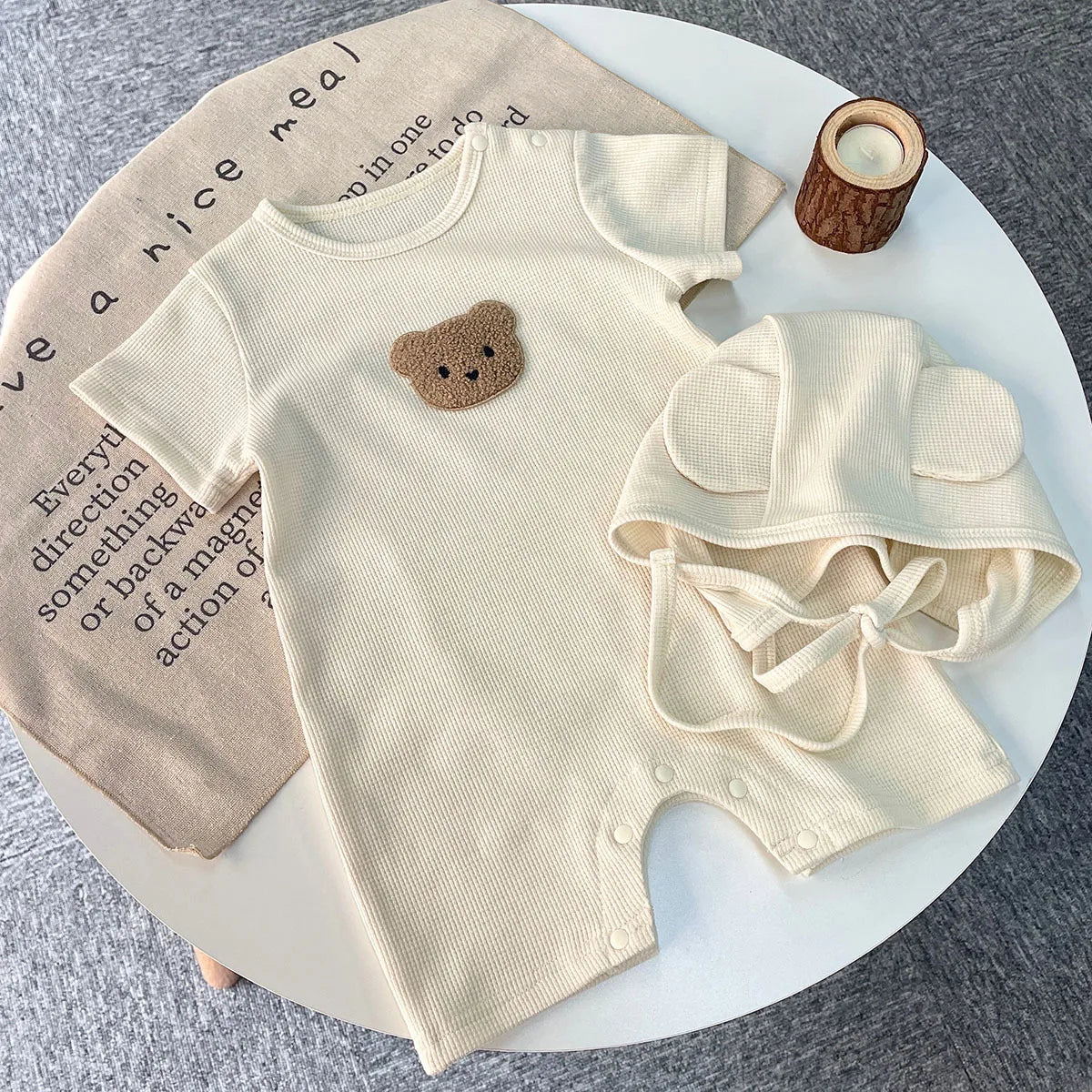 Beige baby romper with a bear face patch and matching hat on a round table. A brown cloth with text and a wood-textured candle holder are nearby.