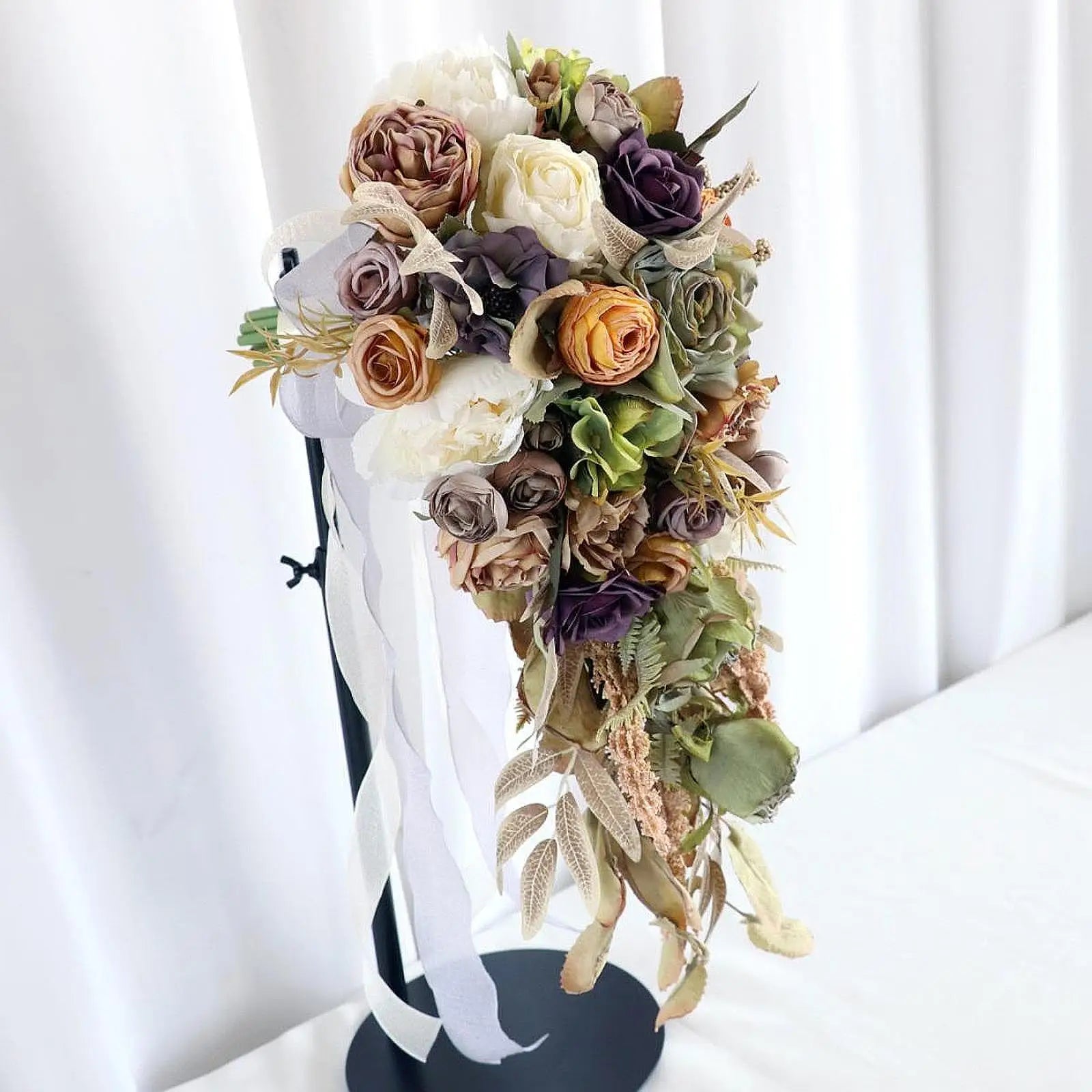 Elegant cascading bouquet with a mix of roses in white, mauve, and peach, accented by greenery and long ribbons, set against a white curtain background.
