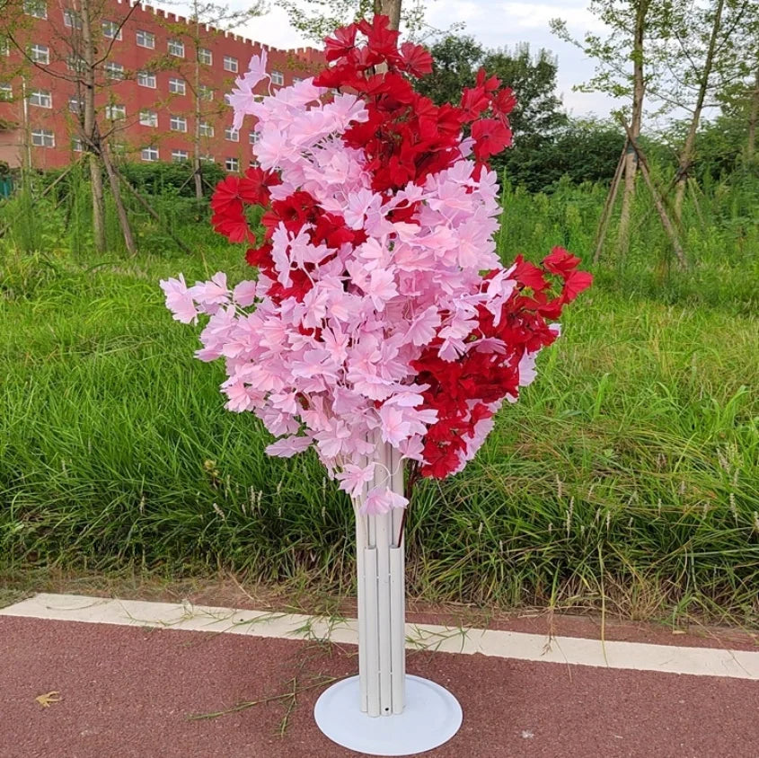 Artificial pink and red flowers on a tall, white stand beside a grassy area. A brick building with multiple windows is visible in the background under a partly cloudy sky.