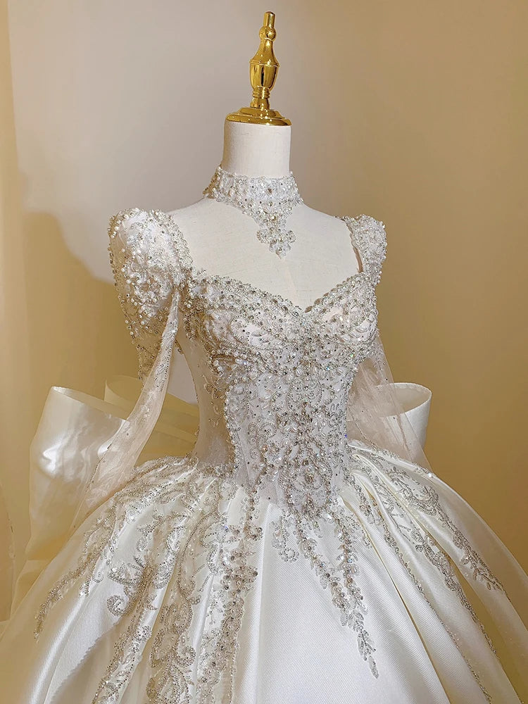 A luxurious white wedding gown on a mannequin, featuring intricate silver beadwork and embroidery. The dress has a full skirt, ornate bodice, and high neckline with embellishments.