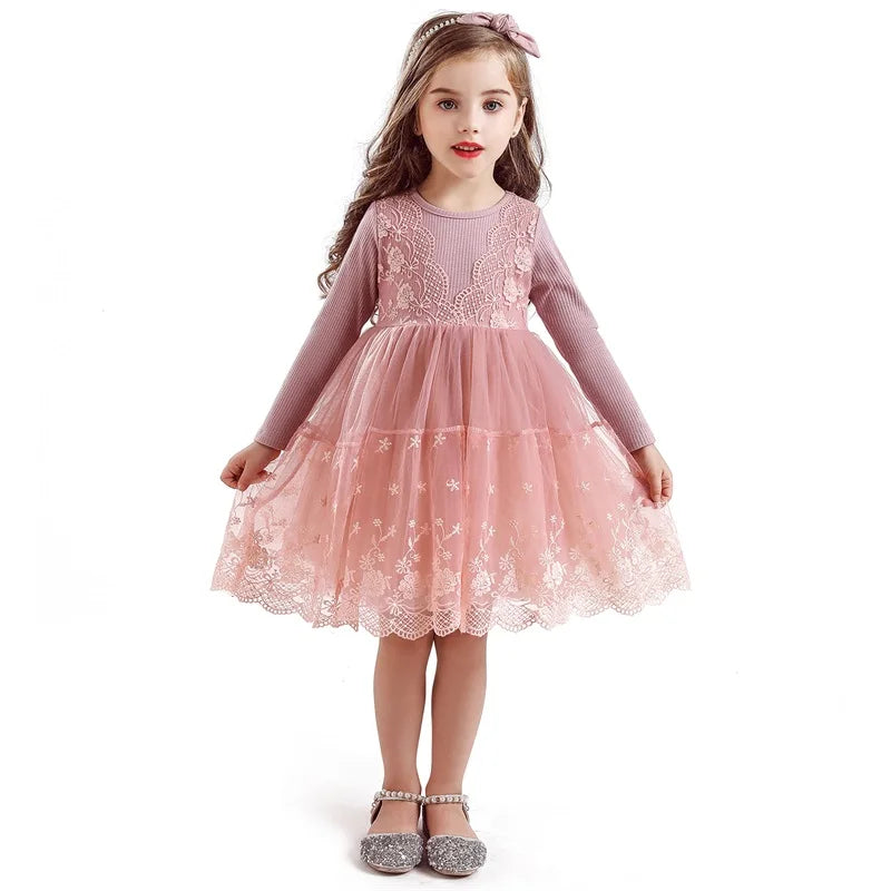 Young girl in a pink lace dress with long sleeves, standing against a white background. She wears a matching headband and silver glittery shoes, with delicate floral patterns on the dress.