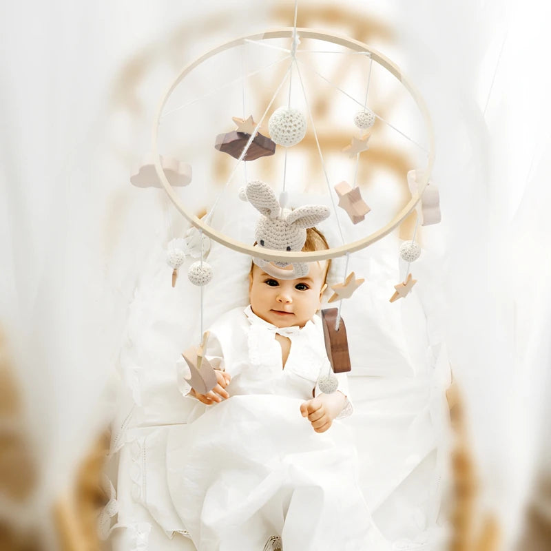 Baby in white outfit lies in crib beneath a decorative mobile with soft toys and shapes. The room is softly lit and the atmosphere is serene and cozy.