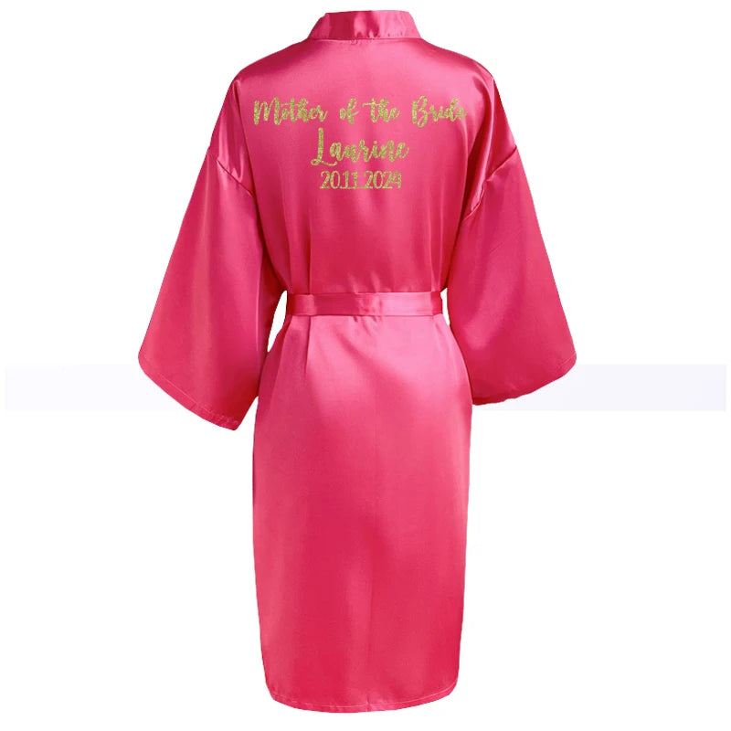 Pink satin robe with text "Mother of the Bride, Laurine, 2024" embroidered in gold on the back. The robe has wide sleeves and a belt, displayed against a white background.