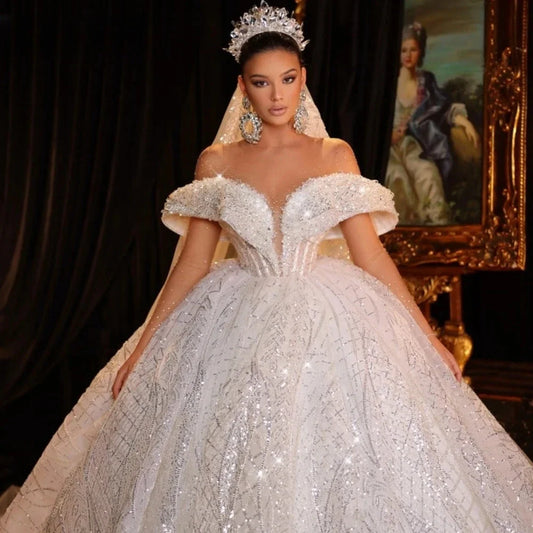 A woman in an ornate off-shoulder wedding gown adorned with sparkling details stands confidently. She wears an elegant tiara and earrings, with a regal portrait behind her.