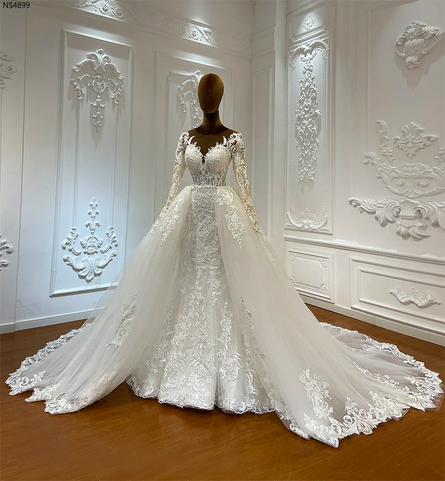 Elegant white wedding dress on a mannequin in an ornate room. The dress features intricate lace details, long sleeves, and a flowing train with floral patterns.