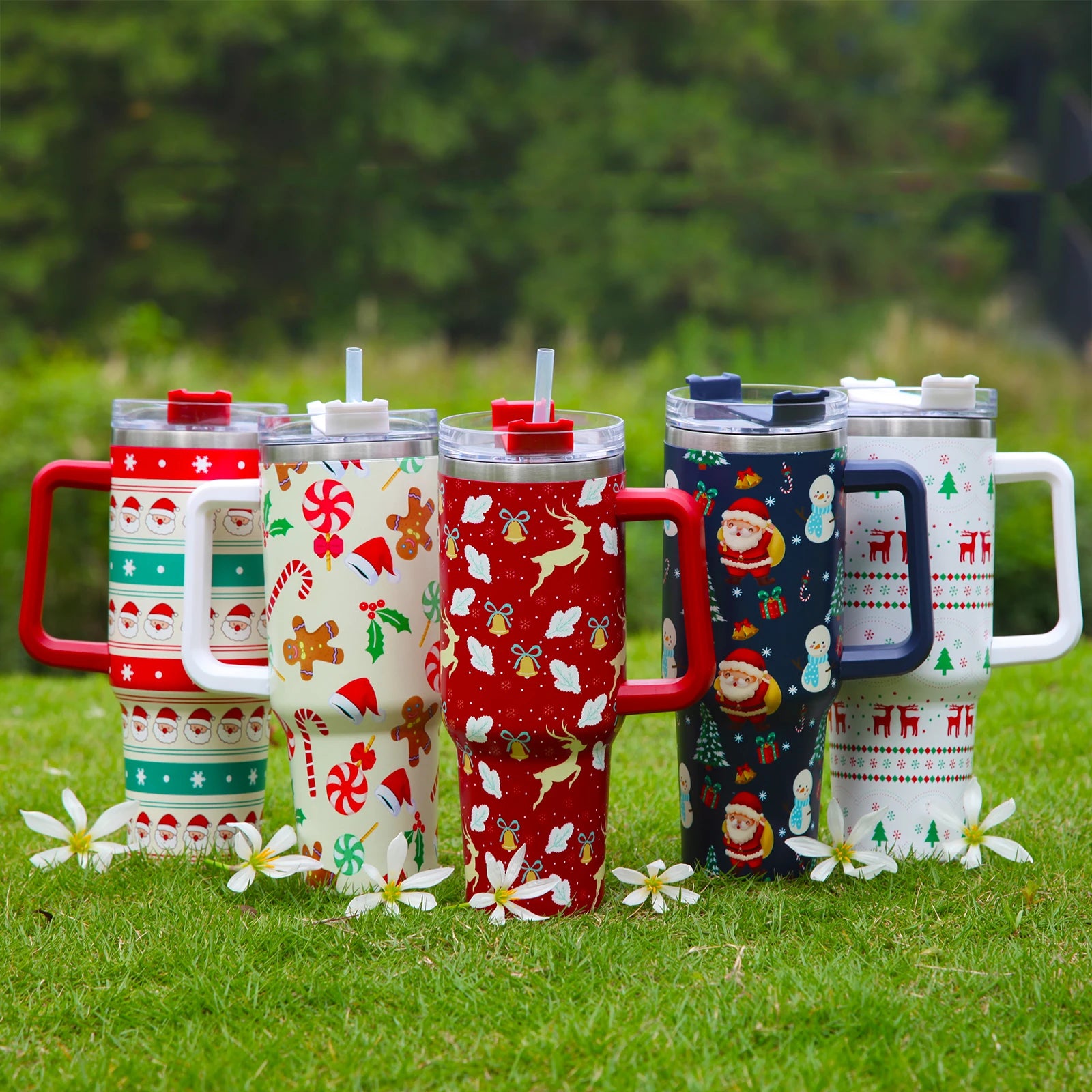 Five festive tumblers with holiday patterns, including Santa, snowmen, and reindeer, arranged on grass with white flowers. Each has a plastic lid and a straw.