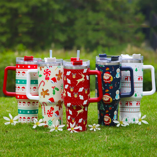 Five festive tumblers with holiday patterns, including Santa, snowmen, and reindeer, arranged on grass with white flowers. Each has a plastic lid and a straw.