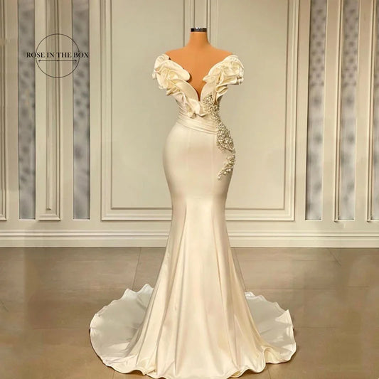 Elegant off-the-shoulder mermaid gown with floral embellishments and intricate ruffles. The dress features a fitted silhouette and a soft, flowing train, set in a formal room.