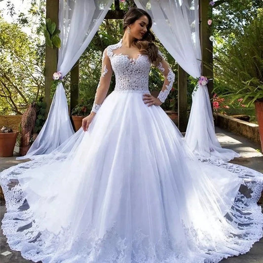Bride in white lace wedding dress with long sleeves stands outdoors. The gown features intricate lace details, a full skirt, and a flowing train, surrounded by lush greenery and draped fabric.