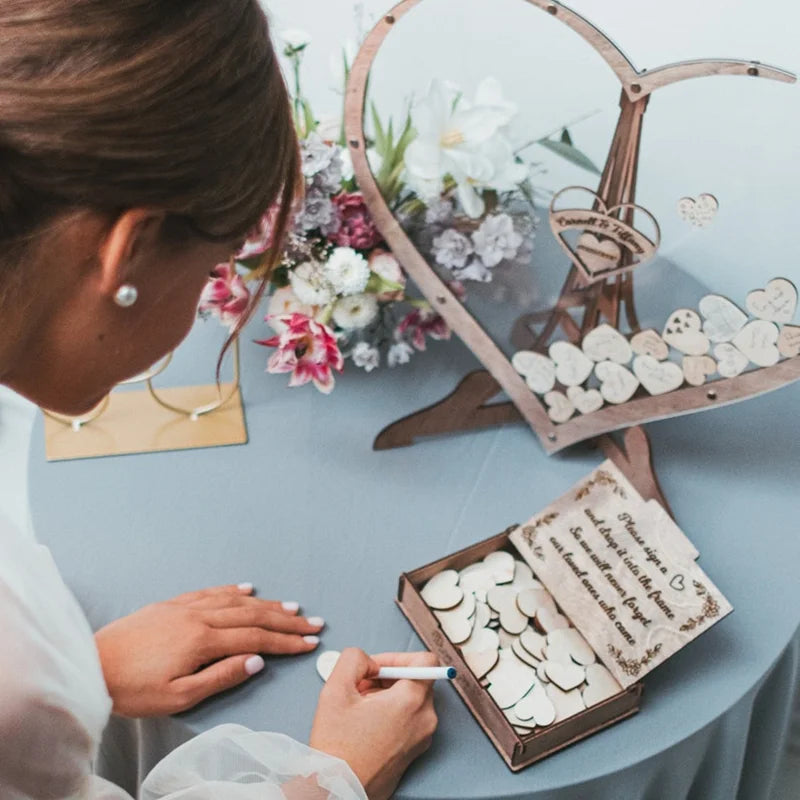 A woman writes on a wooden heart piece, which is part of a wedding guestbook display on a table with flowers. The guestbook is shaped like a heart, filled with signed wooden hearts.