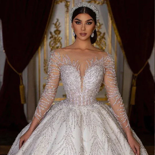 Elegant woman in a shimmering, ornate white gown with intricate beadwork and long sleeves, stands in a richly decorated room with dark curtains and gold accents.