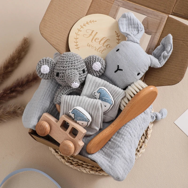 Gift box with baby items including a knitted elephant toy, gray bunny, wooden brush, and toy car, gray socks with whale design, a “Hello World” sign, in a cozy woven basket.