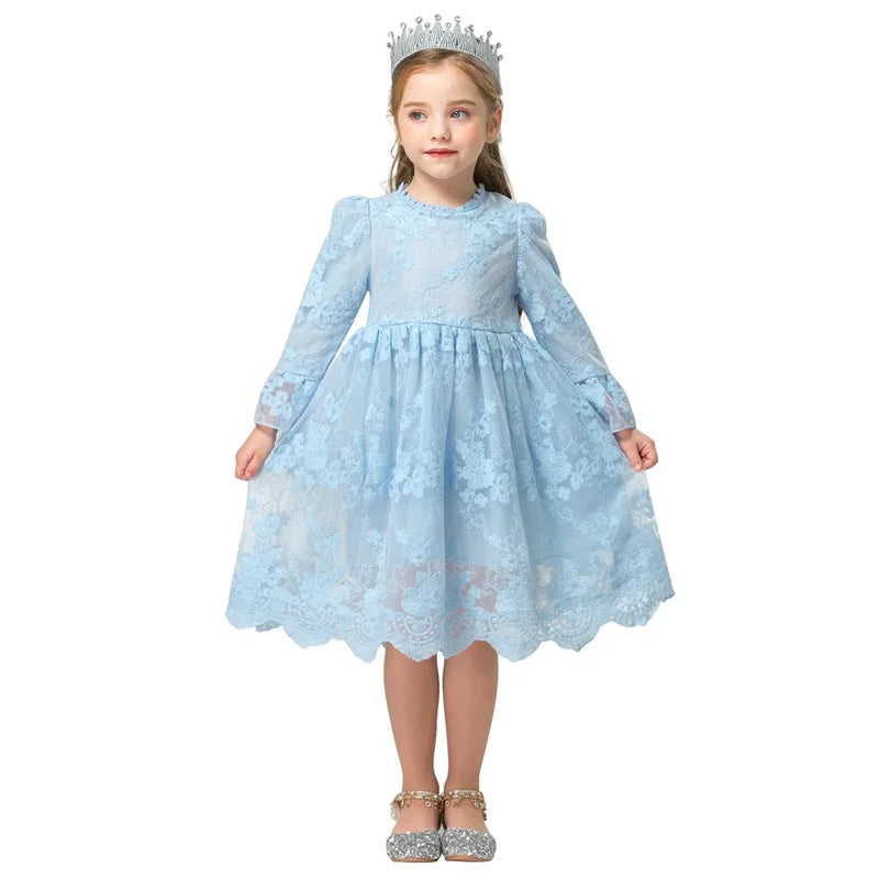 Young girl in a light blue lace dress with puffed sleeves and a tiara, standing with hands holding the dress hem, wearing sparkly silver shoes.