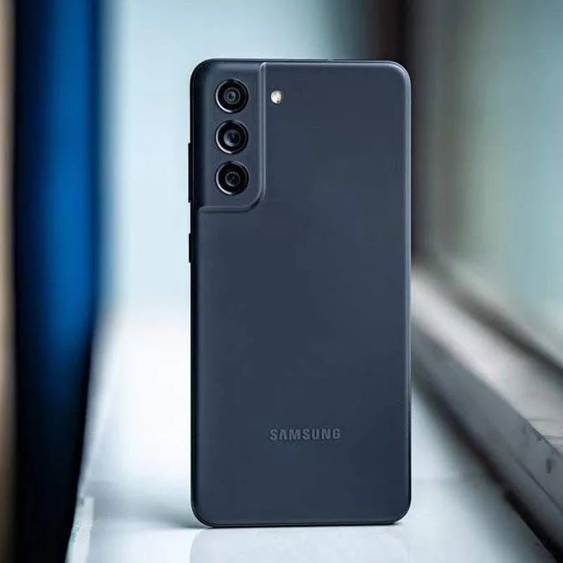 Black Samsung smartphone with a triple camera setup on the upper left. The device is placed on a reflective surface near a window with natural light filtering through.