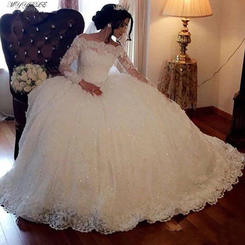 Bride in an elegant, lace off-shoulder ball gown with a sparkling tiara sits on a vintage armchair. A bouquet rests beside her; a lamp on a table softly lights the room.