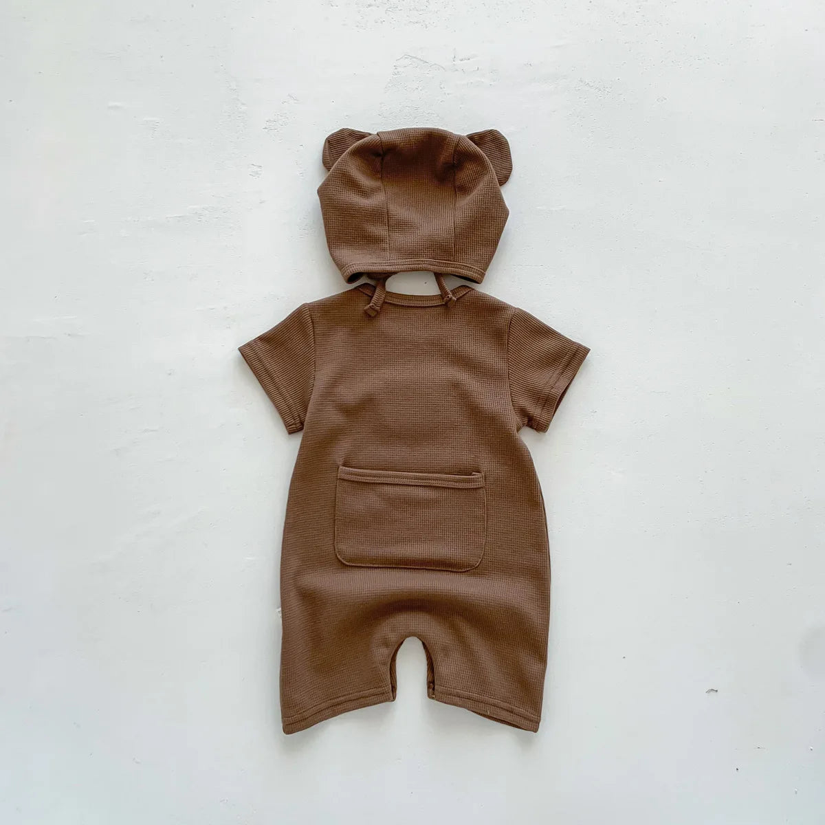 Brown baby romper with short sleeves, a front pocket, and a hood with bear ears, laid flat against a plain white background.