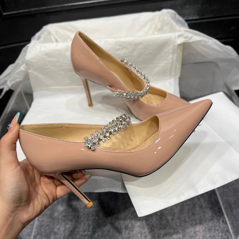 Beige patent leather high heels with pointed toes and rhinestone straps. One shoe is held by a hand, showcasing its elegant design, against a white background.