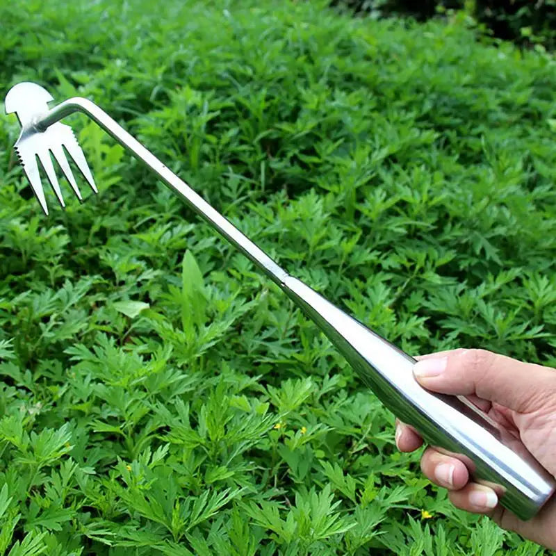 A person holds a stainless steel weeding tool with sharp prongs and an angled tip, poised above green foliage, against a background of dense grass.