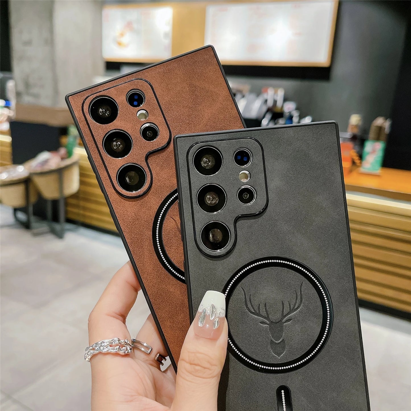 Two smartphones with leather-textured cases, one brown and one black, featuring embossed deer designs and prominent camera arrays. Person with manicured nails holds them. Background: cafe setting.