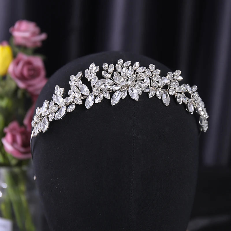 A sparkling silver tiara with intricate floral leaf designs adorned with clear gemstones, displayed on a black velvet stand. Blurred pink and yellow flowers in the background.