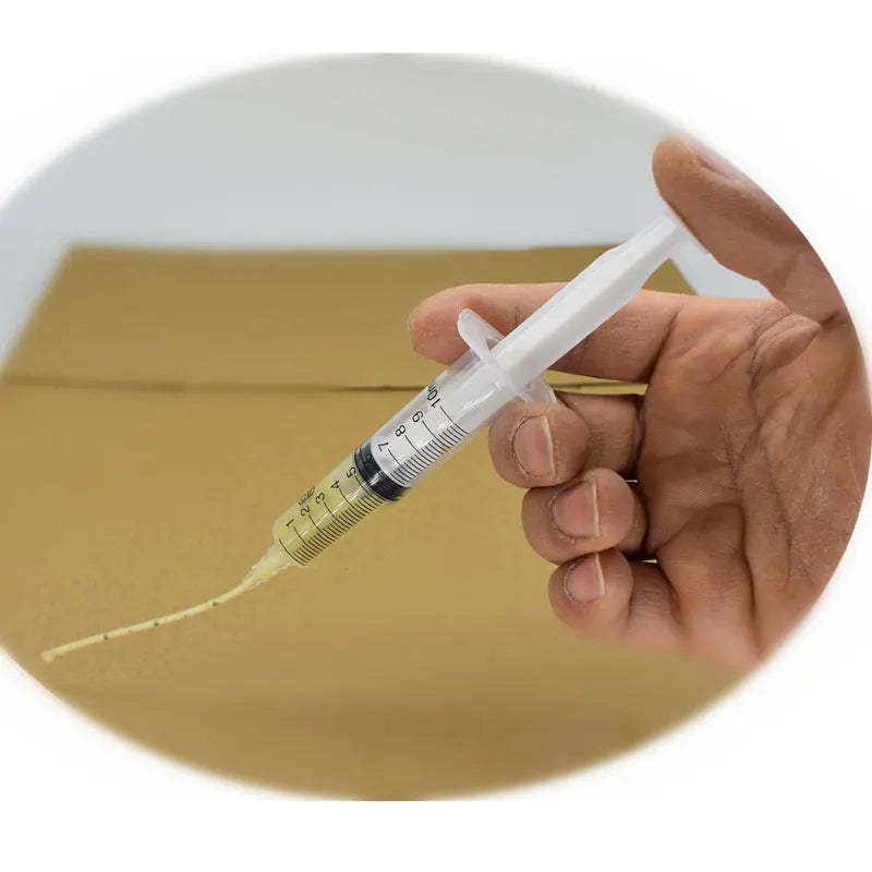 A hand holding a syringe filled with yellow adhesive is squeezing the contents onto a brown surface. The background is out of focus, highlighting the syringe's detailed measurements.