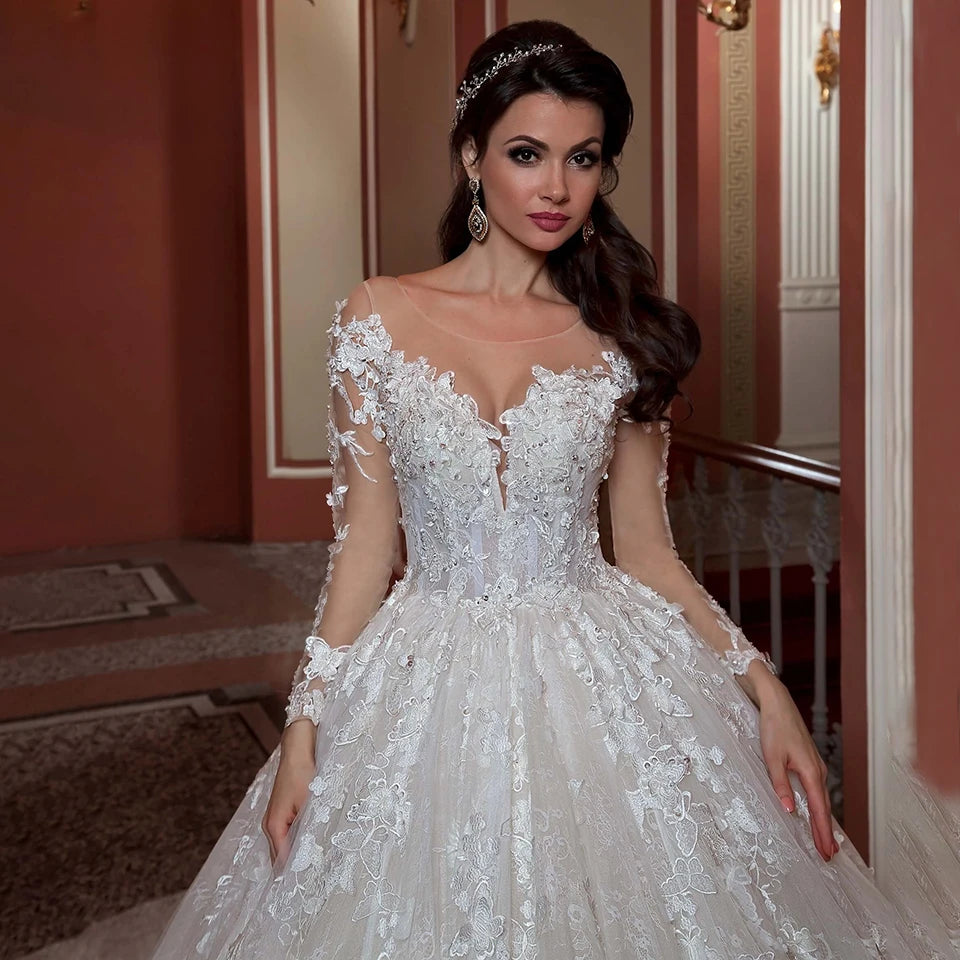A woman in an elegant white lace wedding gown with intricate floral patterns stands in a sophisticated interior, hair styled with a beaded headpiece, and wearing dangling earrings.