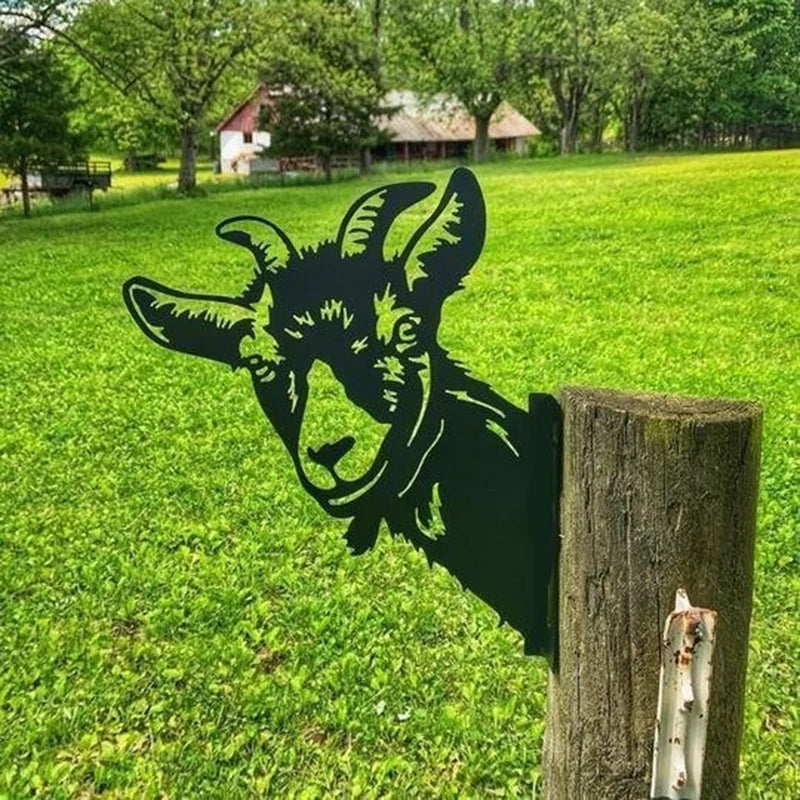 Silhouette of a goat's head attached to a wooden post in a grassy field, with trees and rustic buildings in the background.