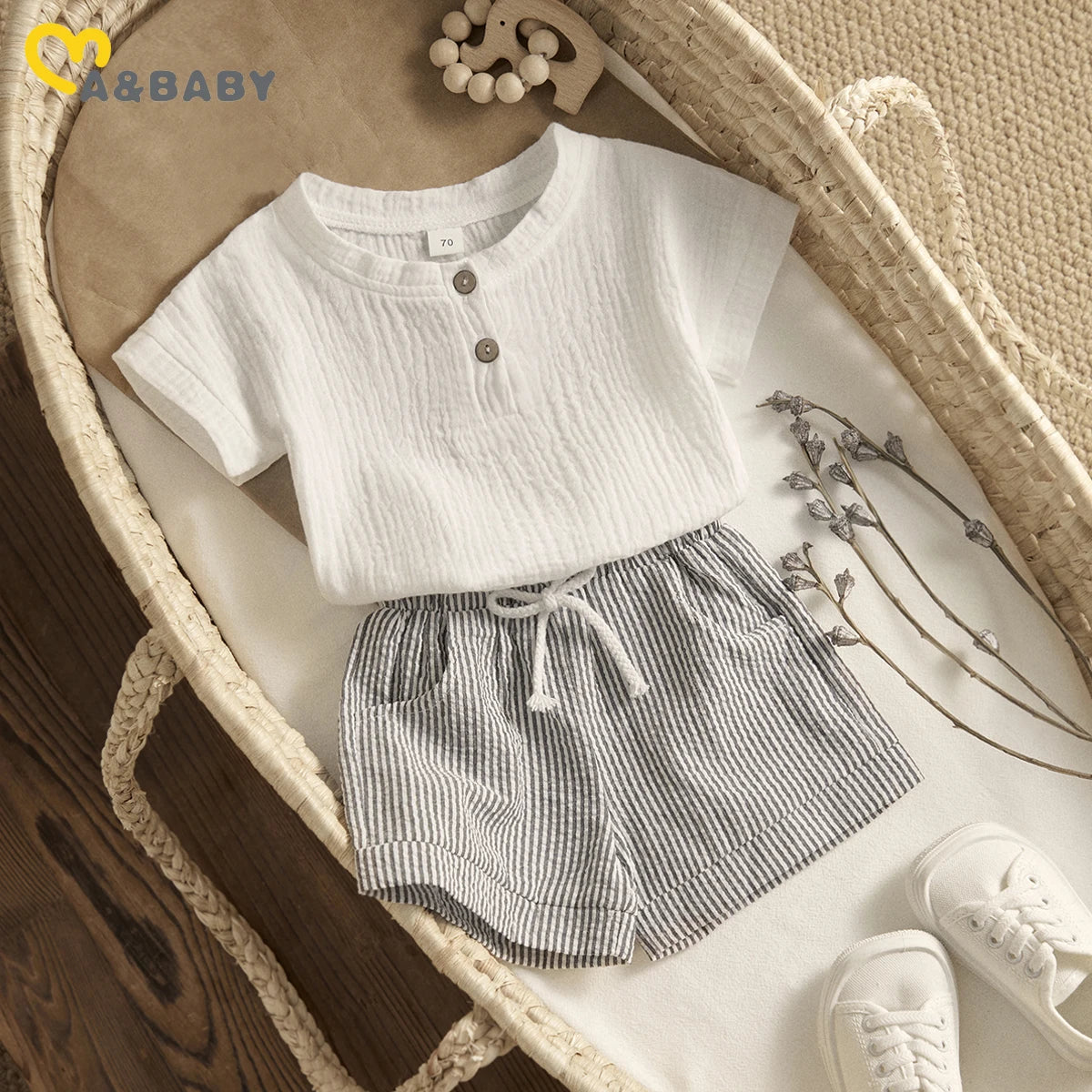 Baby outfit with a white textured shirt, striped shorts, and white shoes, laid out in a woven basket with decorative lavender stems and a wooden toy elephant on brown paper.