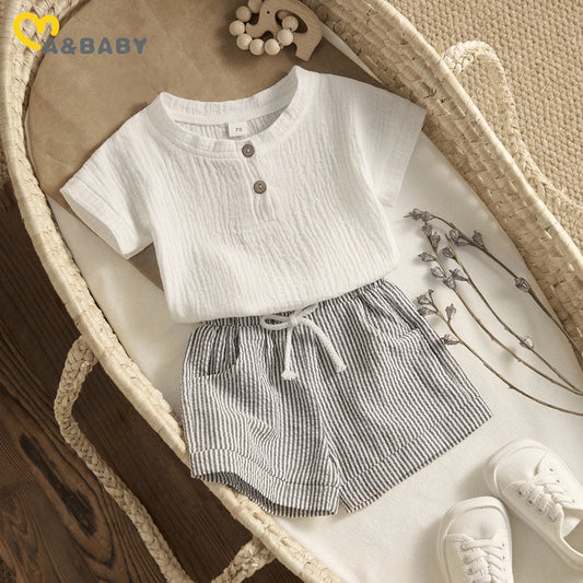 Baby outfit with a white textured shirt, striped shorts, and white shoes, laid out in a woven basket with decorative lavender stems and a wooden toy elephant on brown paper.