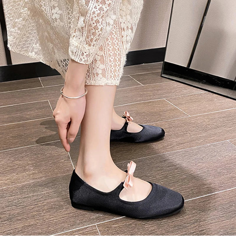 Person in a lace dress adjusts black flats with pink bows on a wood-look tile floor. A silver bracelet is visible on their wrist.