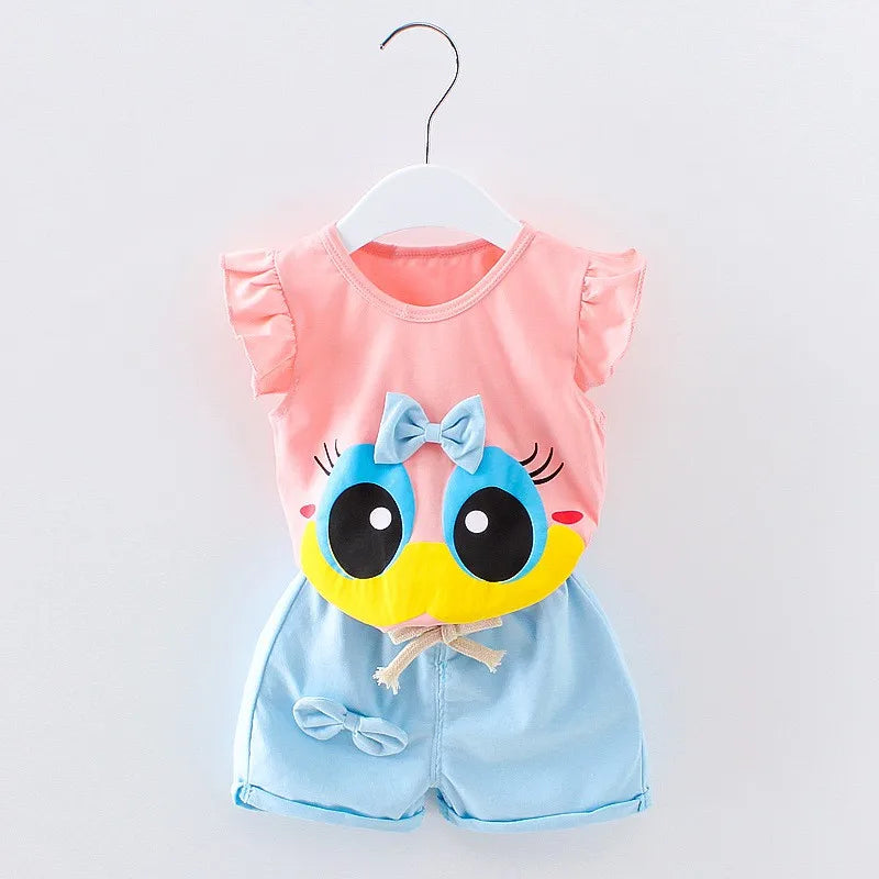 Pink and blue toddler outfit with frilled sleeves, featuring a cartoon face with large eyes and a bow on the chest. The shorts have a drawstring and a small bow accent.
